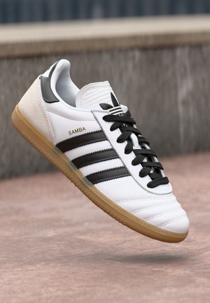 Hvit Adidas Samba-sneaker med svarte striper, svarte lisser, gummisåle og gull "SAMBA"-tekst, satt mot en utendørs steinoverflate som bakgrunn.