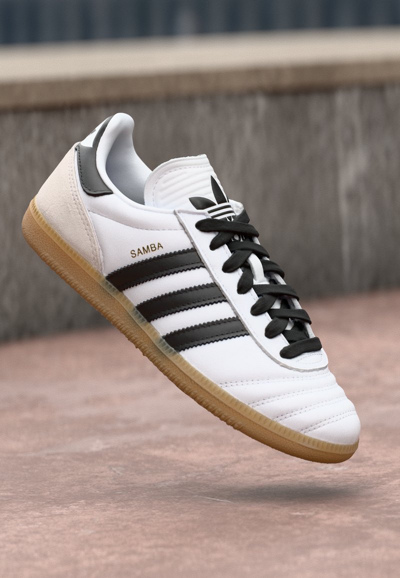 Sapatilha Adidas Samba branca com riscas pretas, atacadores pretos, sola de borracha e texto em dourado "SAMBA", colocada sobre um fundo de superfície de pedra ao ar livre.