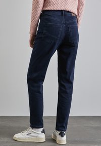 Mörkblå denimjeans med rak skärning, synliga bakfickor och diskreta sömnadsdetaljer. Tillsammans med vita sneakers mot en grå bakgrund.