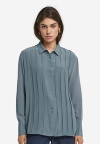 Kvinna med brunt hår som bär guldörringar, en blå veckad skjortblus och mörkblå jeans mot en vit bakgrund.