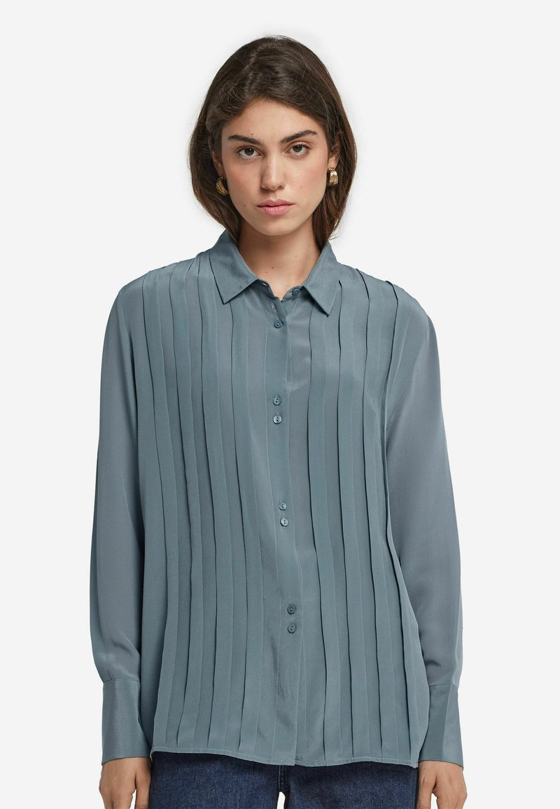 Kvinna med brunt hår som bär guldörringar, en blå veckad skjortblus och mörkblå jeans mot en vit bakgrund.