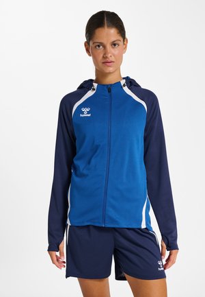 Jonge vrouw met donker haar in een blauw- en marineblauw sportjack met capuchon en bijpassende marineblauwe korte broek, staande tegen een witte achtergrond.