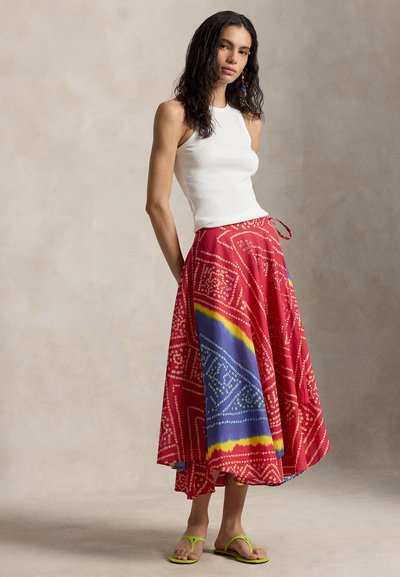 Nainen, jolla on valkoinen hihaton paita, punainen kuvioitu pitkä hame sinisillä ja keltaisilla yksityiskohdilla sekä limenvihreät sandaalit, seisomassa yksivärisen taustan edessä.