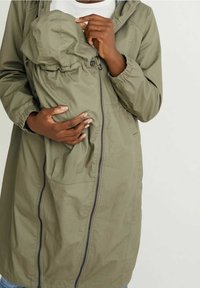 Olivgrön hoodie-jacka med framdragkedja och samlad tyg. Har en ficka för att bära ett barn och elastiska ärmslut. Slät textur.