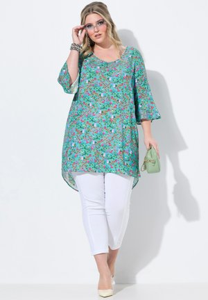 Blumen-Tunika in Türkis, mit kurzen Ärmeln und einem asymmetrischen Saum. Kombiniert mit weißen Hosen und accessoriert mit einer grünen Tasche.
