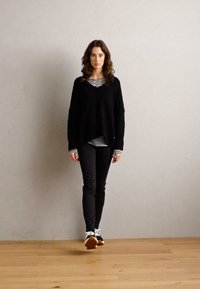 Schwarzer, übergroßer, gestrickter Pullover mit V-Ausschnitt; übereinander getragen mit einem schwarz-weißen gestreiften Hemd; kombiniert mit schwarzen schmalen Hosen und weißen Sneakers.
