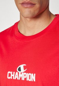 Rød bomulds T-shirt med rund hals, med hvid "CHAMPION" logo og emblem på brystet, glat tekstur, korte ærmer.