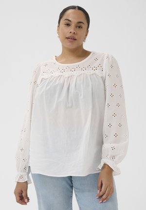 Kobieta o ciemnych włosach, ubrana w biały, dziurkowany top z napuszonymi rękawami i jasnoniebieskie dżinsy, stojąca na tle jednolitego jasnego tła.