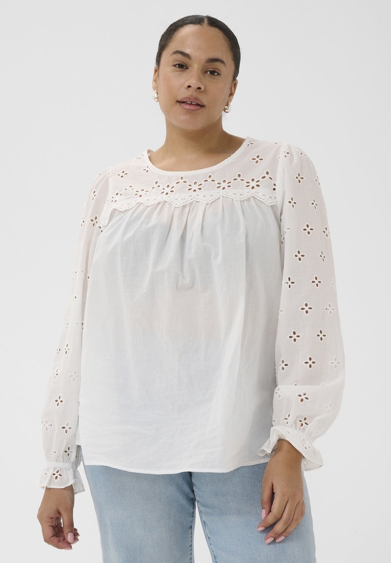 Femme aux cheveux foncés portant un chemisier blanc à œillets avec des manches bouffantes et un jean bleu clair, posant devant un fond uni clair.