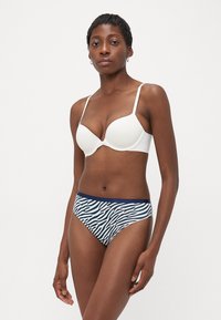 Woman wearing white bra and zebra-print bikini-style underwear standing against plain white background.