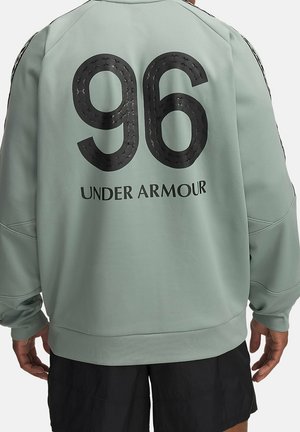 Rückansicht einer Person, die einen hellgrünen Under Armour Pullover mit einer großen schwarzen "96" und dem Markennamen auf dem Rücken trägt, kombiniert mit schwarzen Shorts.
