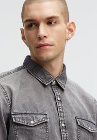 Grau denim Hemd mit durchgehender Knopfleiste, zwei Brusttaschen und klassischem Kragen. Besitzt eine abgenutzte Textur und sichtbare Nähdetails.