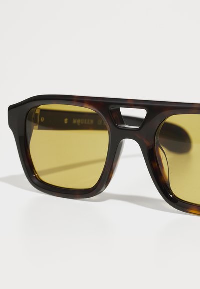 Ochelari de soare din material chel cu lentile dreptunghiulare. Lentile galbene cu nuanță și o decupare distinctă în podul nasului. Ramă din plastic neted, cu hardware minim.