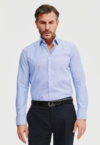 Jasnoniebieska koszula w paski z długimi rękawami, z ko�łnierzykiem zapinanym na guziki i białymi guzikami; zestawiona z ciemnymi spodniami i czarnym paskiem.