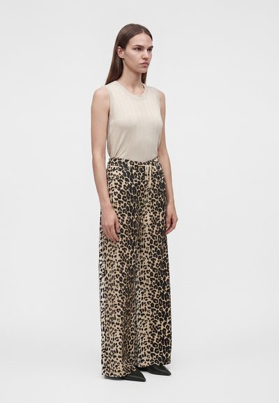 Femme aux longs cheveux bruns portant un top beige sans manches et un pantalon large imprimé léopard avec des chaussures noires pointues.