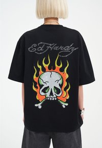 Camiseta negra con un gran gráfico de calavera con acentos verdes, huesos cruzados y llamas vibrantes en naranja y amarillo. "Ed Hardy" está escrito con strass.