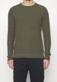 Chemise manches longues vert olive avec encolure ronde, en tissu texturé, présentant des accents cousus le long des poignets et de l'ourlet.