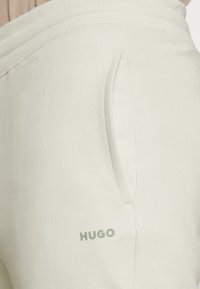 Nahaufnahme einer beigefarbenen Jogginghose mit elastischem Bund und dem "HUGO"-Logo, das nahe der Taschenöffnung aufgedruckt ist.