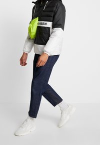 Giacca a vento nera e bianca con cappuccio, dotata di una borsa a tracolla verde brillante. Indossata con jeans scuri e sneakers bianche.