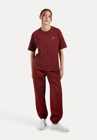 Mörkröd bomull t-shirt och matchande sweatpants, med en avslappnad passform, elastiska manschetter och en liten logotypdetalj på bröstet. Vita sneakers.