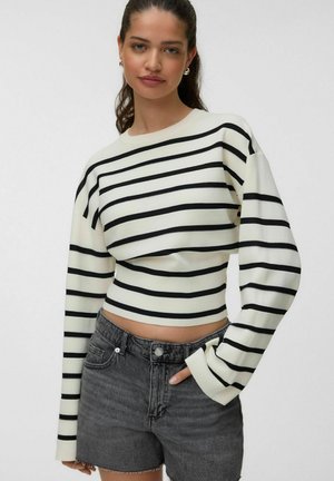 Jeune femme portant un haut blanc à manches longues avec des rayures horizontales noires et un short en jean gris foncé taille haute, posant avec une main dans la poche.