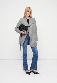 Γκρι γωνιώδες παλτό πάνω από μαύρο τοπ, σε συνδυασμό με μπλε τζιν καμπάνα με μοτίβα και μαύρες μυτερές γόβες, κρατώντας μια μικρή μαύρη τσάντα.