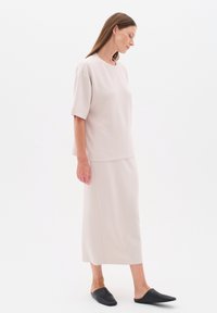 Kvinde iført en løs beige kortærmet top, matchende lang nederdel og sorte slip-on sko stående mod en ensfarvet hvid baggrund.