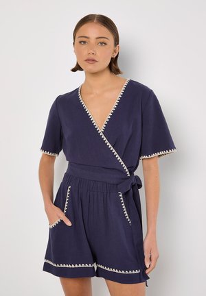 Marineblauer Wickel-Overall mit kurzen Ärmeln, mit cremefarbener gewellter Kante entlang der Ränder und elastischem Taillenbund. Mit seitlichen Taschen.