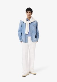 Giacca in denim azzurro chiaro sopra una camicia azzurro chiaro, con pantaloni bianchi e un maglione bianco legato attorno al collo. Sneakers bianche completano il look.