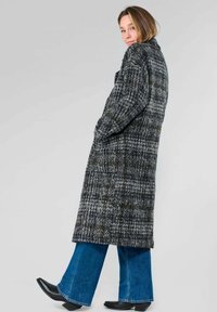 Manteau en pied-de-poule gris et noir avec une coupe longue et ample, doté de poches latérales. Le tissu a un tissage texturé. Porté avec un jean évasé et des bottes.