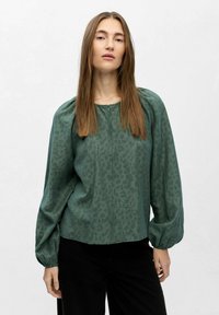 Vrouw met lang steil haar, gekleed in een groene blouse met een patroon en opgezette lange mouwen, en zwarte broek voor een effen lichte achtergrond.