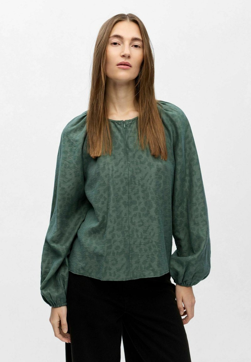 Vrouw met lang steil haar, gekleed in een groene blouse met een patroon en opgezette lange mouwen, en zwarte broek voor een effen lichte achtergrond.