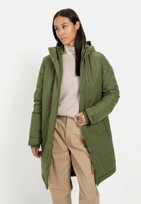 Groene gevoerde parka jas met een capuchon, voorzien van een ritssluiting, zijzakken en een zachte textuur. Draagt over een lichtbeige trui en broek.