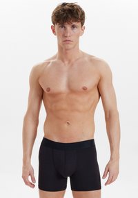Schwarze Baumwoll-Boxershorts mit einem breiten, strukturierten Bund. Der Stoff wirkt glatt und eng anliegend und hat ein Design mit einer Fronttasche.