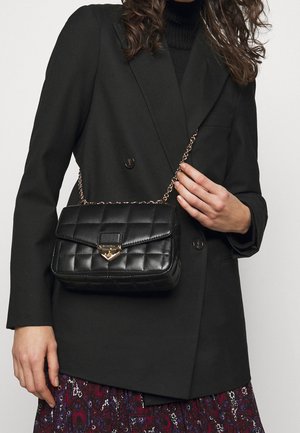 Femme en blazer noir à double boutonnage et jupe à motifs tenant un petit sac bandoulière noir matelassé avec une chaîne dorée.