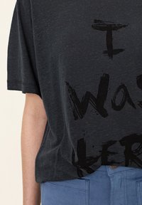 Tmavě šedé texturované tričko s krátkým rukávem a černým potiskem "I WAS HERE" na přední straně. Nosené s modrými kalhotami s detailem na knoflík.