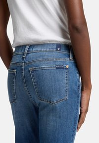 Jeans en denim bleu foncé avec une taille mi-haute, des poches arrière cousues et une petite étiquette de marque sur la taille avec des garnitures en métal doré.