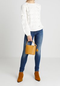 Personne portant un pull en maille blanc, un jean skinny bleu, des bottines marron, tenant un petit sac à main marron par la poignée.