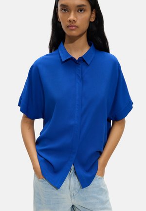 Młoda osoba ubrana w luźną, krótką, królewsko niebieską koszulę zapinaną na guziki i jasnoniebieskie dżinsy z rękami w kieszeniach na jednolitym tle.