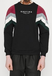Persona que lleva una sudadera negra con rayas en Chevron rojas, grises, blancas y verdes en las mangas y el texto "KAOTIKO SINCE 1999" en el pecho.