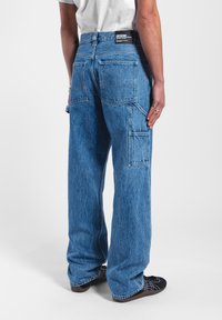 Jeans de mezclilla azul de pierna ancha con dos bolsillos traseros y un bolsillo tipo cargo lateral. La tela tiene un acabado ligeramente texturizado. Etiqueta de la marca negra visible en la cintura.
