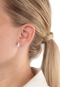 Primo piano di un orecchio di donna con piccolo orecchino a cerchio d'argento, capelli biondi raccolti e parte di un colletto bianco visibile.