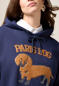 Granatowy hoodie z kapturem, ozdobiony grafiką pluszowego, brązowego psa z akcentami z cyrkonii oraz haftowanym napisem "PARIS DOG" powyżej.
