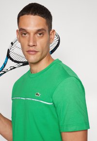 Groene katoenen t-shirt met een ronde hals, een witte bies over de borst en een klein logo. Een tennisracket staat er achter.