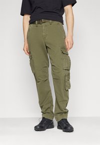 Olivgrüne Cargo-Hose mit mehreren Seitentaschen, Gürtelschlaufen und einer lockeren Passform, kombiniert mit schwarzen Schuhen.