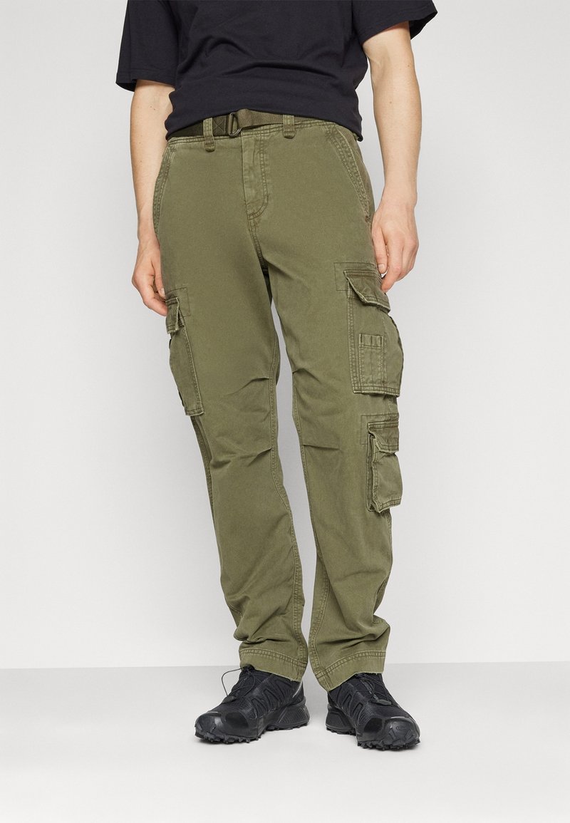 Olivgrüne Cargo-Hose mit mehreren Seitentaschen, Gürtelschlaufen und einer lockeren Passform, kombiniert mit schwarzen Schuhen.