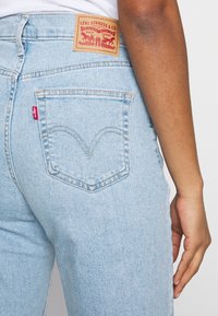 Ljusblå denimjeans med en distinkt bakfickdesign och röd etikett, med en beige läderlapp med varumärkeslogotyp och text.