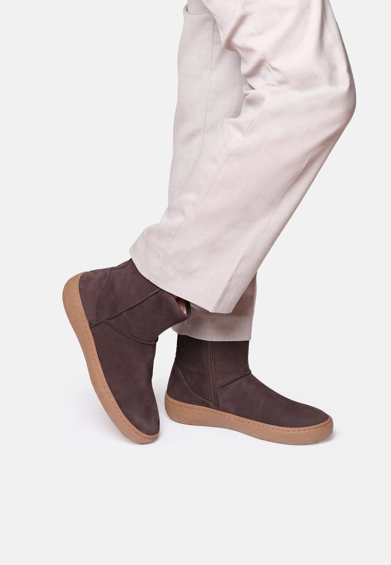Bottines en daim marron avec une semelle en caoutchouc beige, dotées d'une fermeture éclair sur le côté et de coutures minimales, portées avec un pantalon de couleur claire.