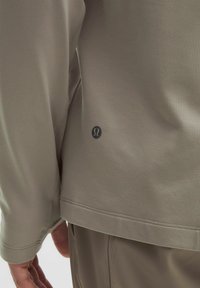 Cămașă cu mânecă lungă, de culoare taupe deschis, dintr-un material neted, având un tiv curbat și un logo mic în apropierea tivului de jos.