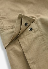 Beige stof med en rutenetmønster, en folded krave og mørke metal snapknapper. Glat tekstur og elegante syninger synlige.